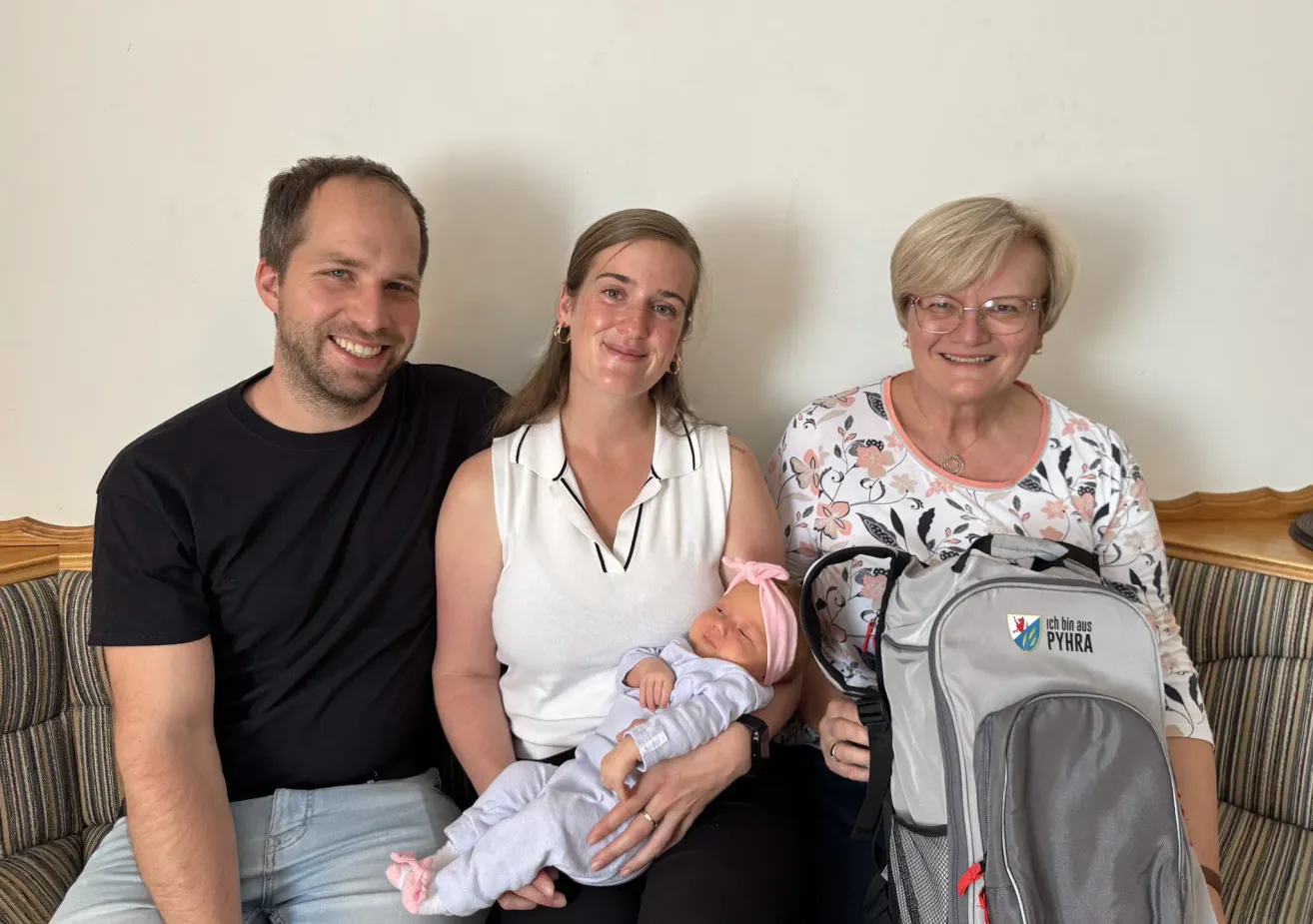Three people are smiling and posing for a picture with a baby on a woman's lap. The baby is wearing a pink headband.