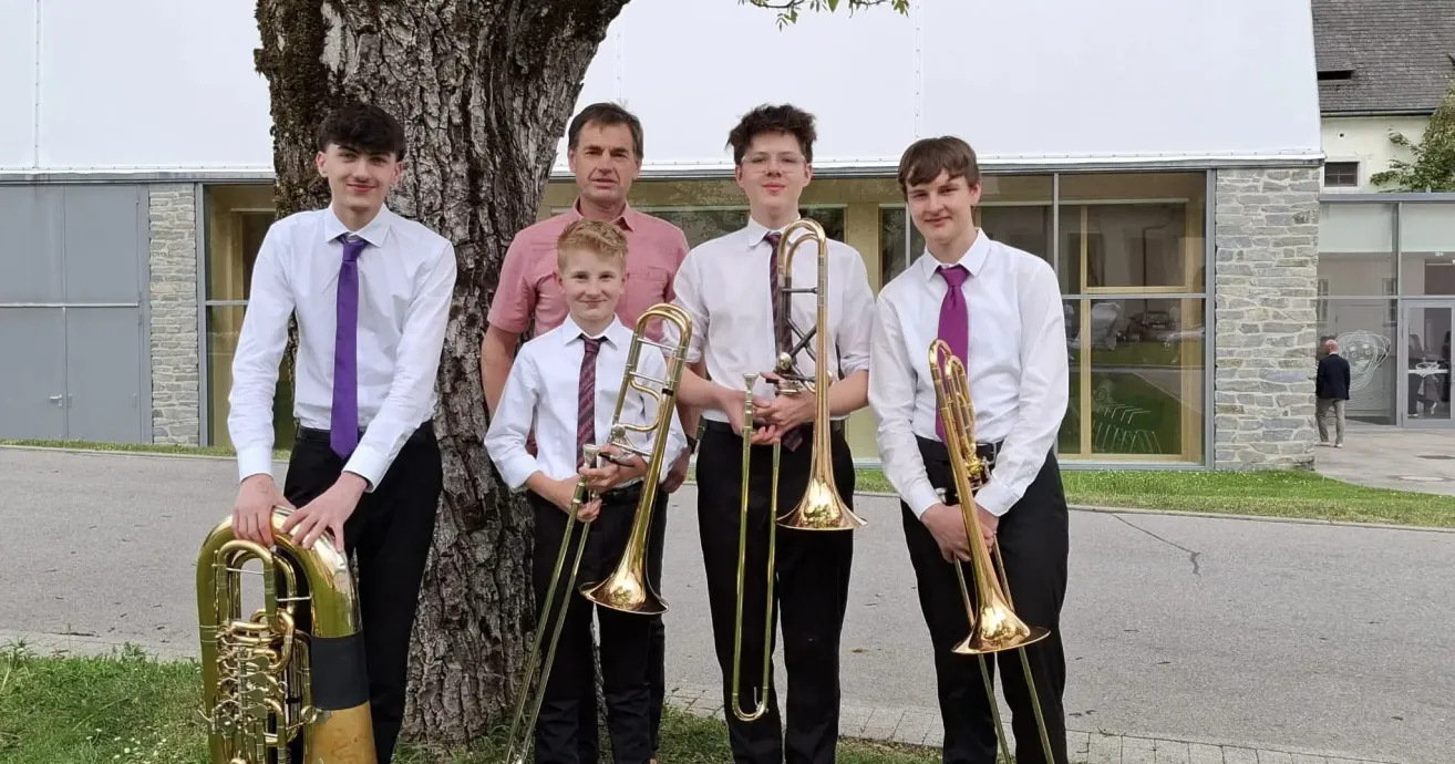 A group of young musicians stands outside, holding trombones. They are dressed in white shirts and ties. A tree trunk is behind them, and a building is visible in the background.