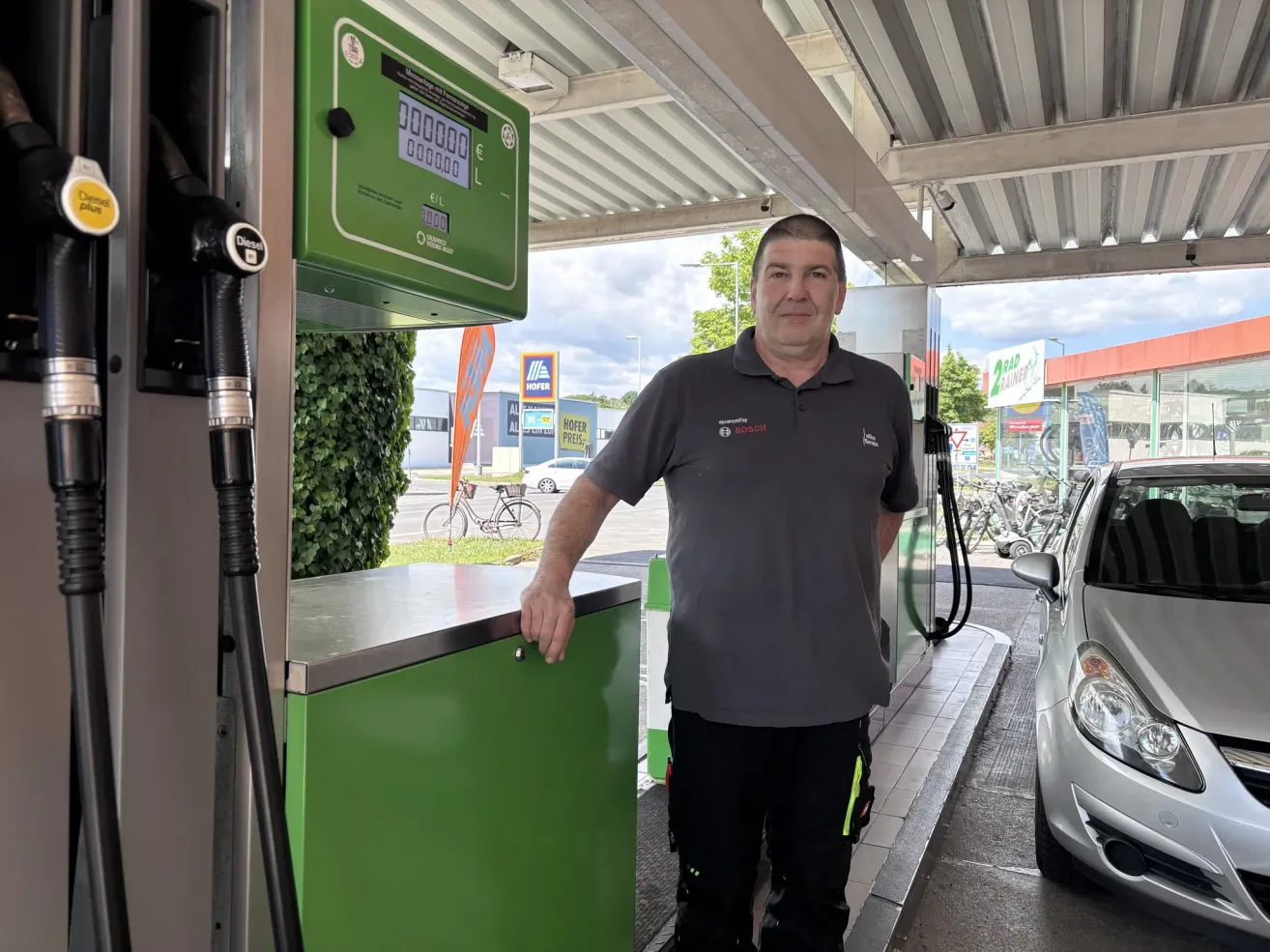 A man in a gray shirt and black pants is standing at a gas pump with a green sign, posing for a picture.
