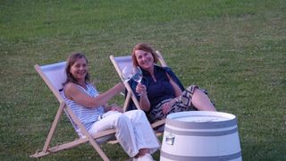 Two women sit in folding chairs on grass, smiling. They hold wine glasses. One woman wears a sleeveless shirt, the other a blouse. A wooden barrel is nearby.