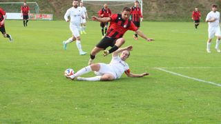 A soccer match with players in white and red jerseys. One player jumps to block the ball, while another player slides on the grass.