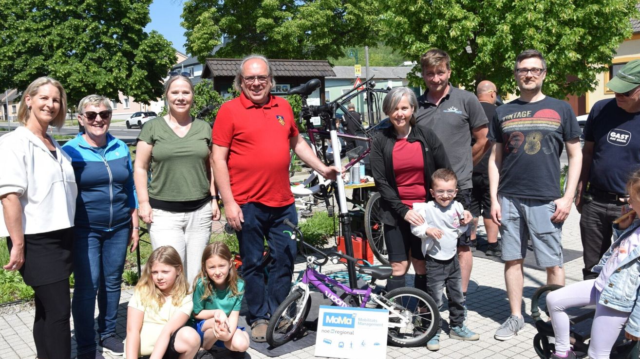 Eine Familie steht mit einem Kind und zwei Fahrrädern in einem Park mit Bäumen und Leuten im Hintergrund. Der Mann im roten Shirt hält ein Schild von Mobilitas Management.