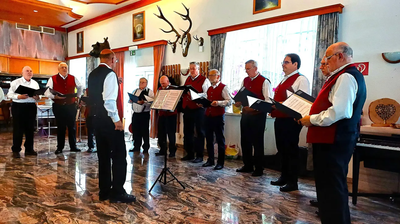 Ein Chor von Männern in roten Weste und schwarzen Hosen singt in einem Raum mit Marmorböden. Ein offenes Buch liegt auf einem Ständer vor ihnen.