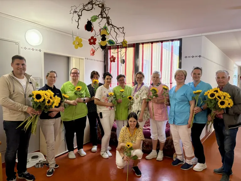 Eine Gruppe von Frauen in einheitlichen Kleidungsstücken steht in einem Raum mit Sonnenblumen, lächelt und hält Sträuße. Sie stehen vor einer weißen Wand mit einem Fenster und dekorativen Aufhängungen.