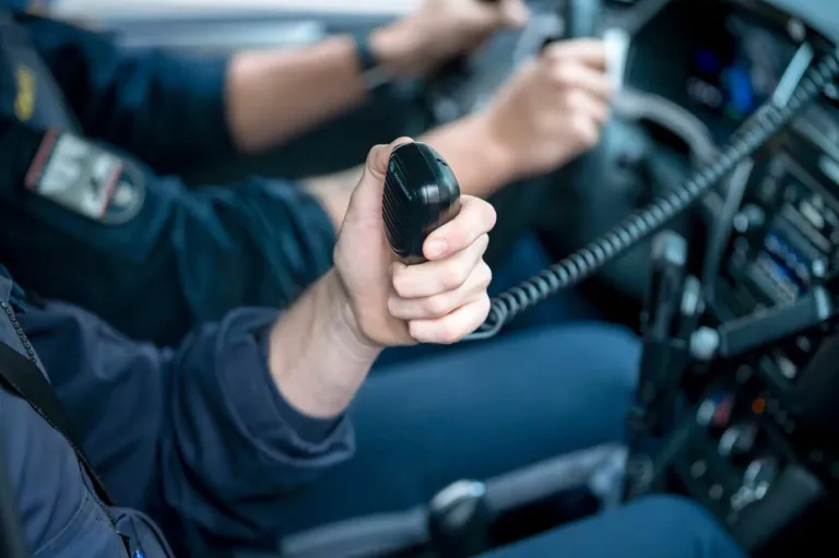 Nahaufnahme einer Hand, die ein schwarzes Gerät in einem Fahrzeuginnenraum hält. Eine Person mit einer Armbanduhr fährt.