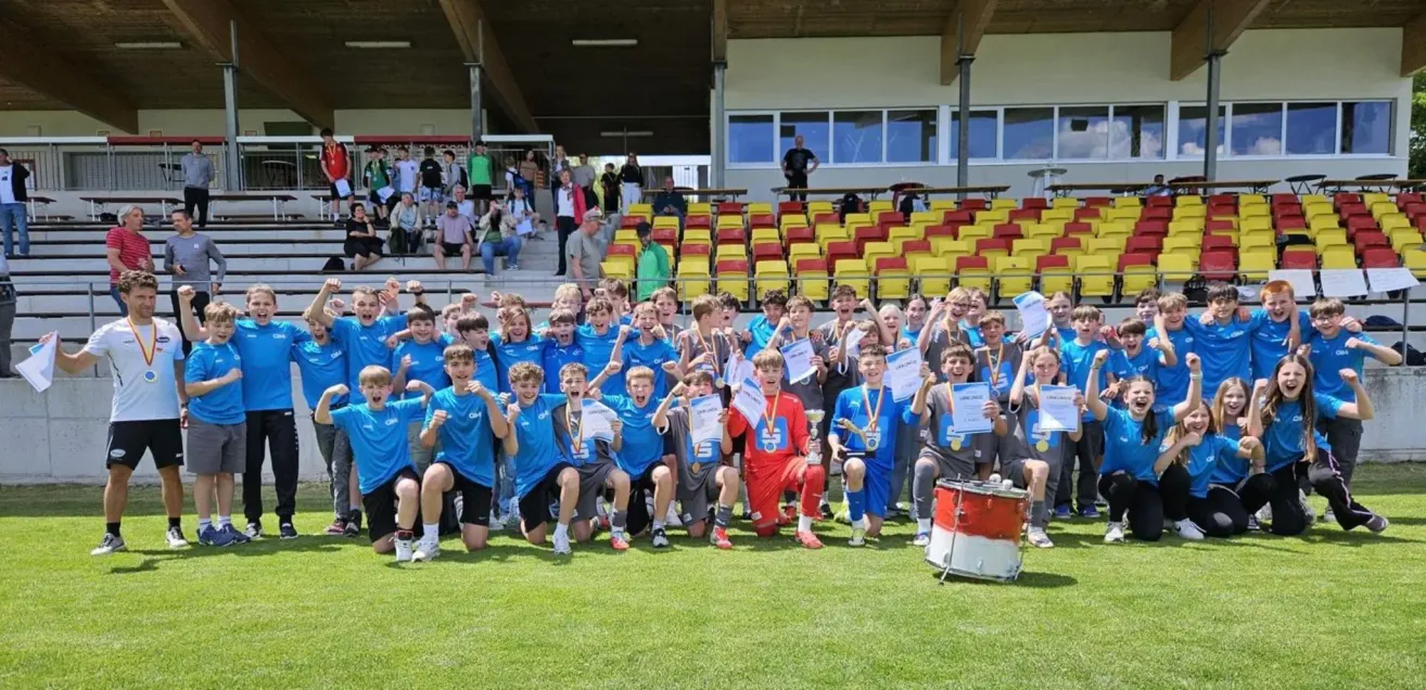 Eine Gruppe junger Athleten in blauen Uniformen mit Medaillen um den Hals posiert für ein Foto auf einem Rasenfeld. Dahinter sind leere Tribünen und einige Menschen stehen auf den Stufen.