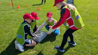 Eine Gruppe von Kindern in grünen Westen und Mützen arbeitet mit einem Erwachsenen, der Papiere in der Hand hält, auf einem Rasenfeld mit Verkehrskegeln.