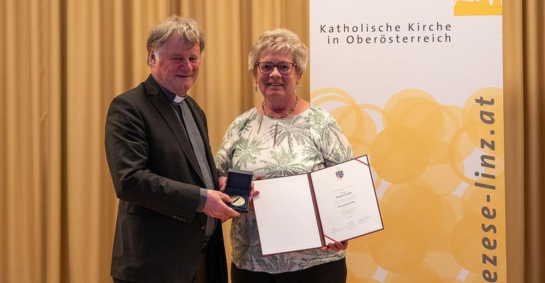 Ein Mann und eine Frau stehen lächelnd vor einer Kulisse mit einem Zertifikat in der Hand. Der Mann trägt einen schwarzen Anzug und eine Krawatte, die Frau ein Blumenhemd. Hinter ihnen befindet sich eine gelbe Kulisse mit dem Text 'Katholische Kirche in Oberosterreich'.