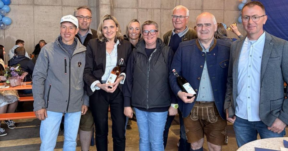 Gruppe von Menschen posiert für ein Foto mit Weinflaschen in der Hand, zwei Frauen vorn lächeln. Ein Mann rechts in traditioneller bayerischer Kleidung, andere lässig gekleidet.