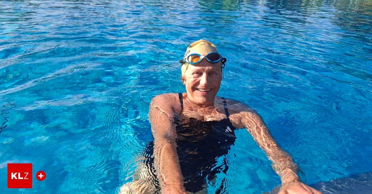 Eine ältere Frau schwimmt in einem Pool mit Schwimmbrille und Schwimmmütze und lächelt in die Kamera. Sie trägt einen schwarzen Badeanzug. Das Wasser ist klar und blau.