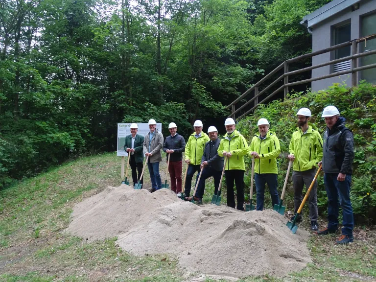 Eine Gruppe von Männern in gelben Jacken und weißen Schutzhelmen hält Schaufeln in einem Grasbereich in der Nähe einer Erdaufschüttung.