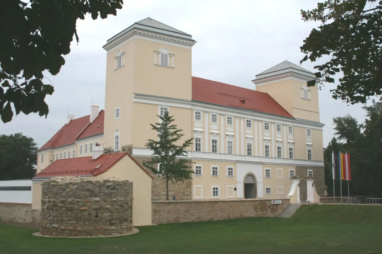 An old historical building with a red roof and many windows, surrounded by a stone wall. A tree stands in front of the building.