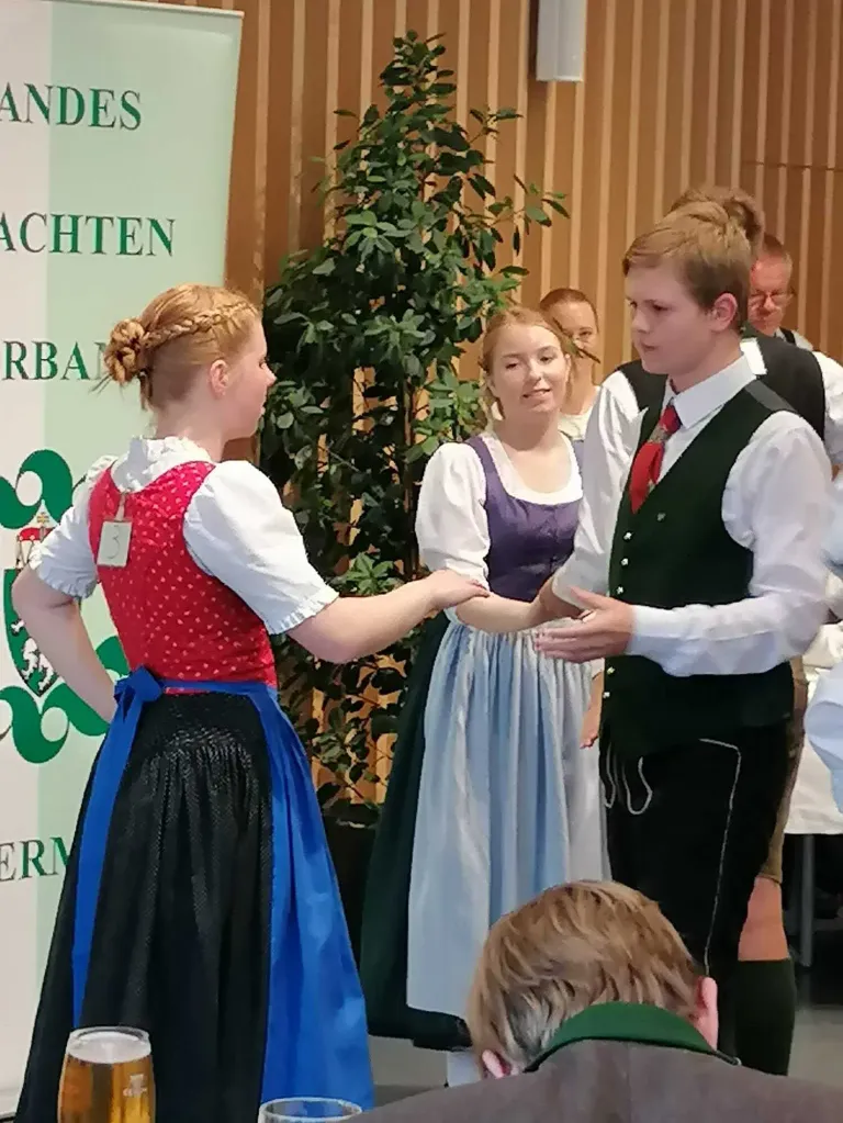 A group of people in traditional clothing are standing. Two young people are talking to each other. Behind them is a plant and a banner.