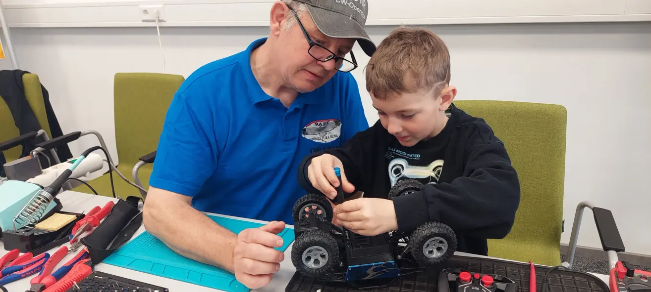A man in glasses and a blue shirt with a logo assists a boy with a screwdriver on a black toy truck.