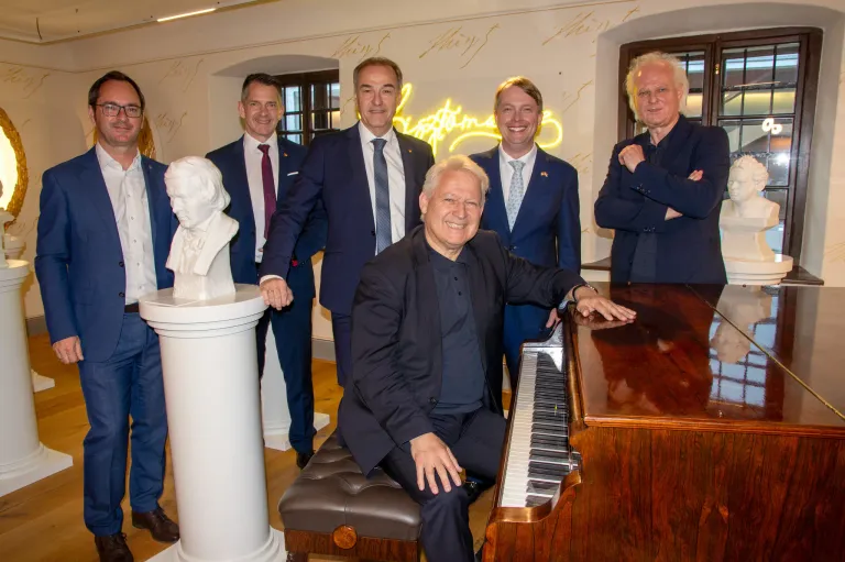 Fünf Männer stehen in einem Raum, einer sitzt an einem Klavier und lächelt, mit einer Büste von Beethoven hinter ihm. Die anderen stehen, alle in Anzügen, einer mit verschränkten Armen.