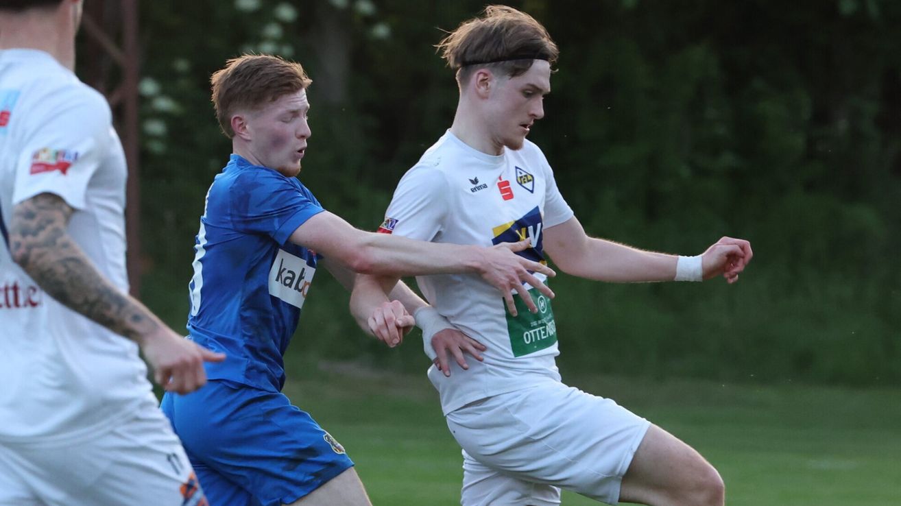 Zwei junge Männer spielen Fußball auf einem Feld. Einer trägt ein blaues Trikot, der andere ein weißes. Der weiße Spieler versucht, den blauen Spieler zu blocken.