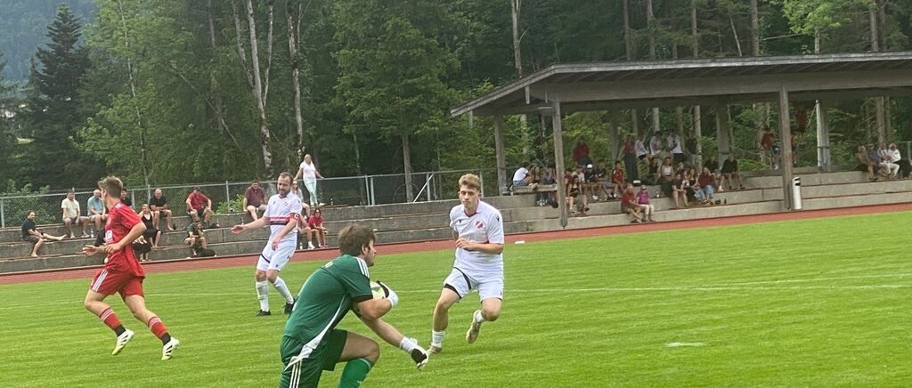 Fußballspieler sind auf dem Feld, ein Torwart bereit, einen Schuss zu blockieren. Zuschauer schauen von den Tribünen zu.