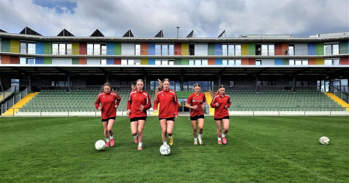 Six female soccer players run on a field, each dribbling a ball. An empty stadium with colorful buildings stands behind them.