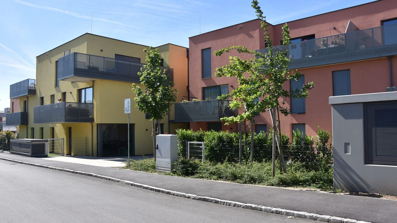Zwei moderne Wohnungen mit Balkonen, eine gelb und eine rosa, stehen neben einer Straße mit Gehweg und Bäumen. Ein kleiner Versorgungskasten befindet sich in der Nähe des Bordsteins.