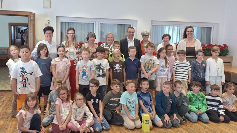 A group of children and adults pose for a photo, with some wearing glasses and sneakers. One adult holds a bouquet of flowers. A yellow box labeled NO is on the floor.