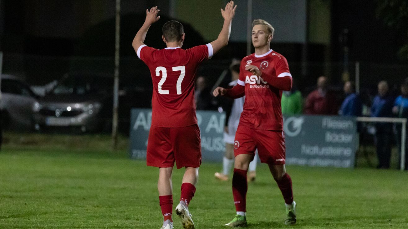 Two soccer players, wearing red uniforms, celebrate on the field with hands raised. One wears jersey number 27, the other has a red shirt with 'ASVi' and a logo. A third player and spectators are in the background.