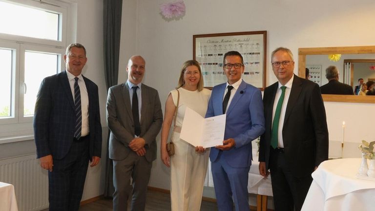 Fünf Menschen stehen in einem Raum, einer hält ein Zertifikat. Sie sind in formeller Kleidung gekleidet. Der Mann rechts trägt eine grüne Krawatte.