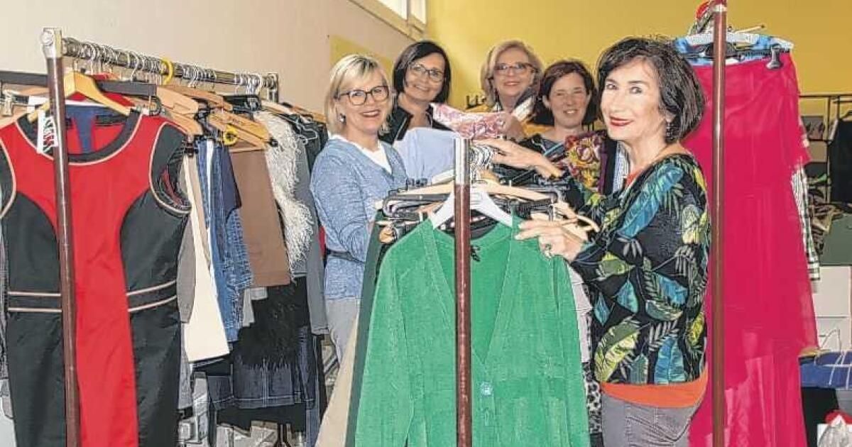 Fünf Frauen stehen lächelnd hinter einem Kleiderständer. Jede Frau hält oder untersucht ein Kleidungsstück und zeigt verschiedene Stile. Der Ständer ist mit verschiedenfarbigen und gestylten Kleidungsstücken gefüllt.