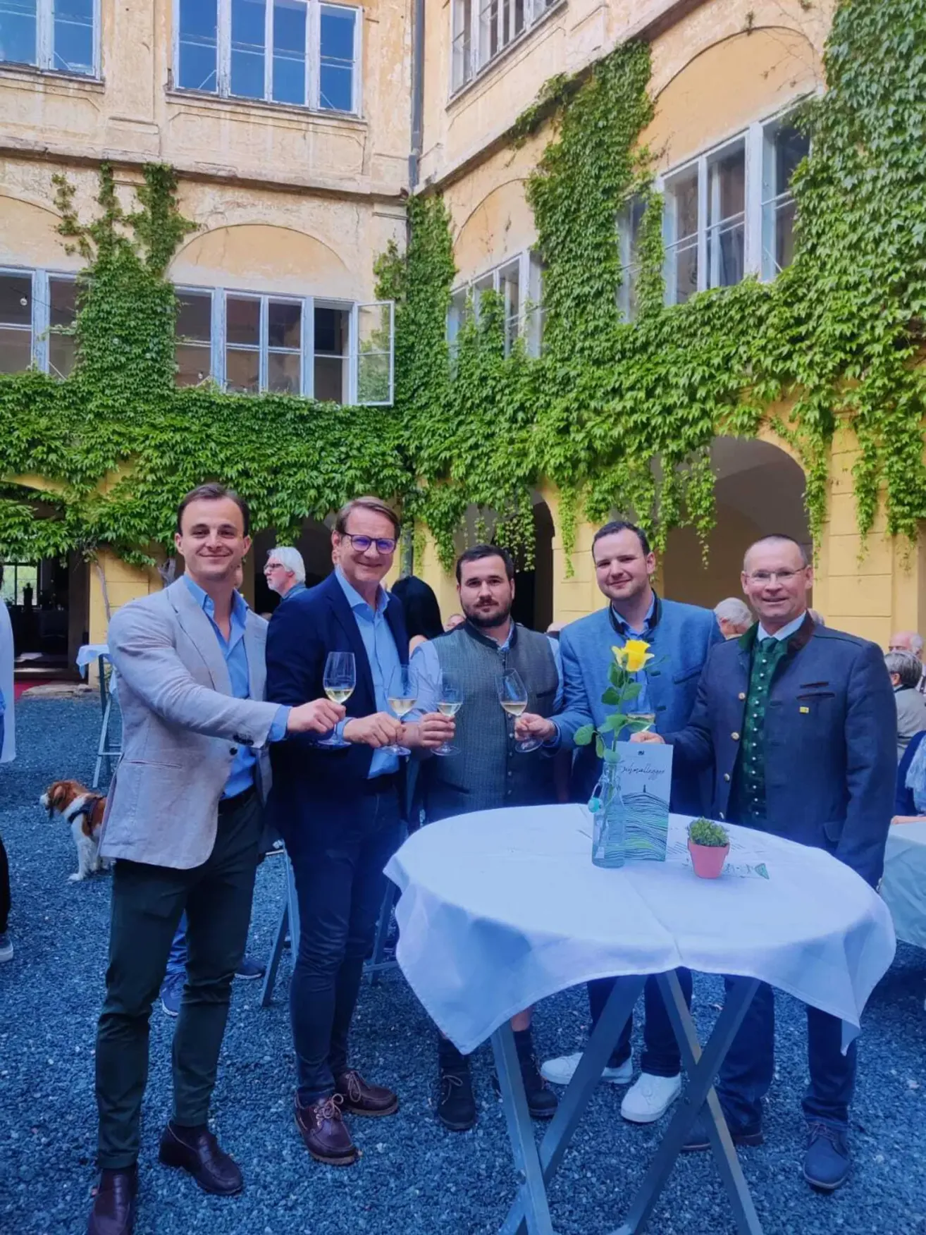Five men, all dressed in suits, are smiling and holding wine glasses outside. A table with a flower vase is in front of them. Behind them is a yellow building with a dog in the background.