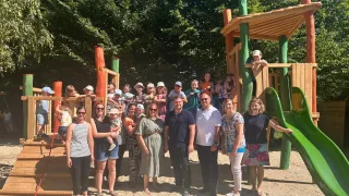 Eine Gruppe von Erwachsenen und Kindern posiert auf einem Spielplatz. Einige stehen vor einem hölzernen Spielhaus, während andere in der Nähe einer grünen Rutsche sind. Bäume spenden im Hintergrund Schatten.