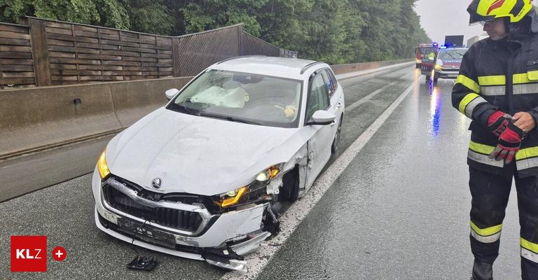 Ein weißer Sedan ist auf dem Seitenstreifen einer Autobahn beschädigt. Die Front ist schwer beschädigt, mit ausgelösten Airbags. Ein Rettungsfahrzeug kommt von der gegenüberliegenden Spur.