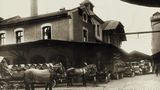 Ein Schwarz-Weiß-Foto zeigt Pferde, die Wagen vor einer Fabrik ziehen. Fässer und andere Waren werden entladen.