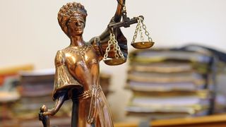 Eine bronzene Statue der blinden Lady Justice hält eine Waage und ein Schwert. Dahinter befindet sich ein Stapel Papiere.