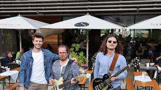 Drei Männer stehen in einem Restaurant im Freien und posieren mit ihren Gitarren. Hinter ihnen sind ein Tisch und Stühle arrangiert. Einer der Männer trägt eine Brille und eine Jeansjacke, während ein anderer lange Haare und ein blaues Hemd trägt. Eine Pflanze und Sonnenschirme sind ebenfalls vorhanden.