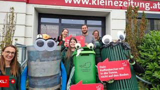 Eine Gruppe von Menschen in Kostümen steht vor einem Gebäude mit Mülleimern. Ein Schild sagt 'Dein Kaffeebecher will in die Tonne, nicht auf den Boden.'