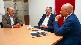 Drei Männer in Anzügen sitzen um einen Tisch in einem Büro und führen ein Gespräch. Auf dem Tisch stehen eine Tasse mit Untertasse und eine Broschüre.