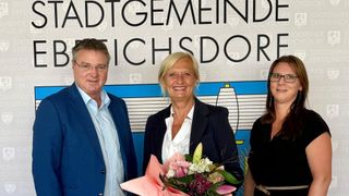 Zwei Erwachsene, ein Mann und eine Frau, stehen lächelnd vor einer Kulisse. Die Frau hält einen Blumenstrauß in der Hand, während der Mann direkt in die Kamera schaut. Eine dritte Person ist teilweise rechts sichtbar.