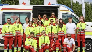 Eine Gruppe von Notfallsanitätern in Neon-Uniformen posiert für ein Foto vor ihrem Krankenwagen.