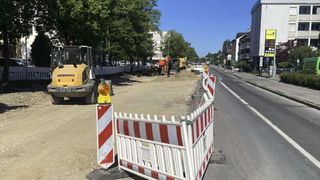 Eine Straße im Bau, mit einer rot-weißen Absperrung, einem großen Bagger und Gebäuden im Hintergrund.