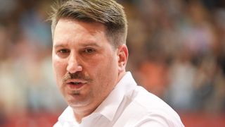 Ein Mann mit Schnurrbart und Bart, der ein weißes Hemd trägt, sieht fokussiert aus. Er hat kurze, gestylte Haare und einen ernsten Gesichtsausdruck. Der Hintergrund ist unscharf, möglicherweise ein Innenraum.