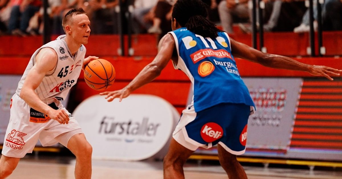Zwei Basketballspieler spielen auf dem Platz. Ein Spieler dribbelt den Ball, während der andere versucht, ihn zu blocken. Der Platz hat eine rote Bank im Hintergrund.