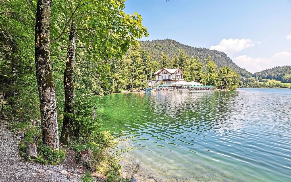 Ein ruhiger See umgeben von üppiger Grünfläche, ein malerisches Haus mit Terrasse und einem Berg im Hintergrund.