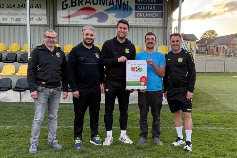 Fünf Männer stehen auf einem Rasenfeld. Einer hält ein Plakat. Sie tragen Sportbekleidung und Turnschuhe. Hinter ihnen zeigt ein Gebäude ein Schild mit dem Namen G.BRAUMANN und der Aufschrift Heizung Sanitär Brunnen.