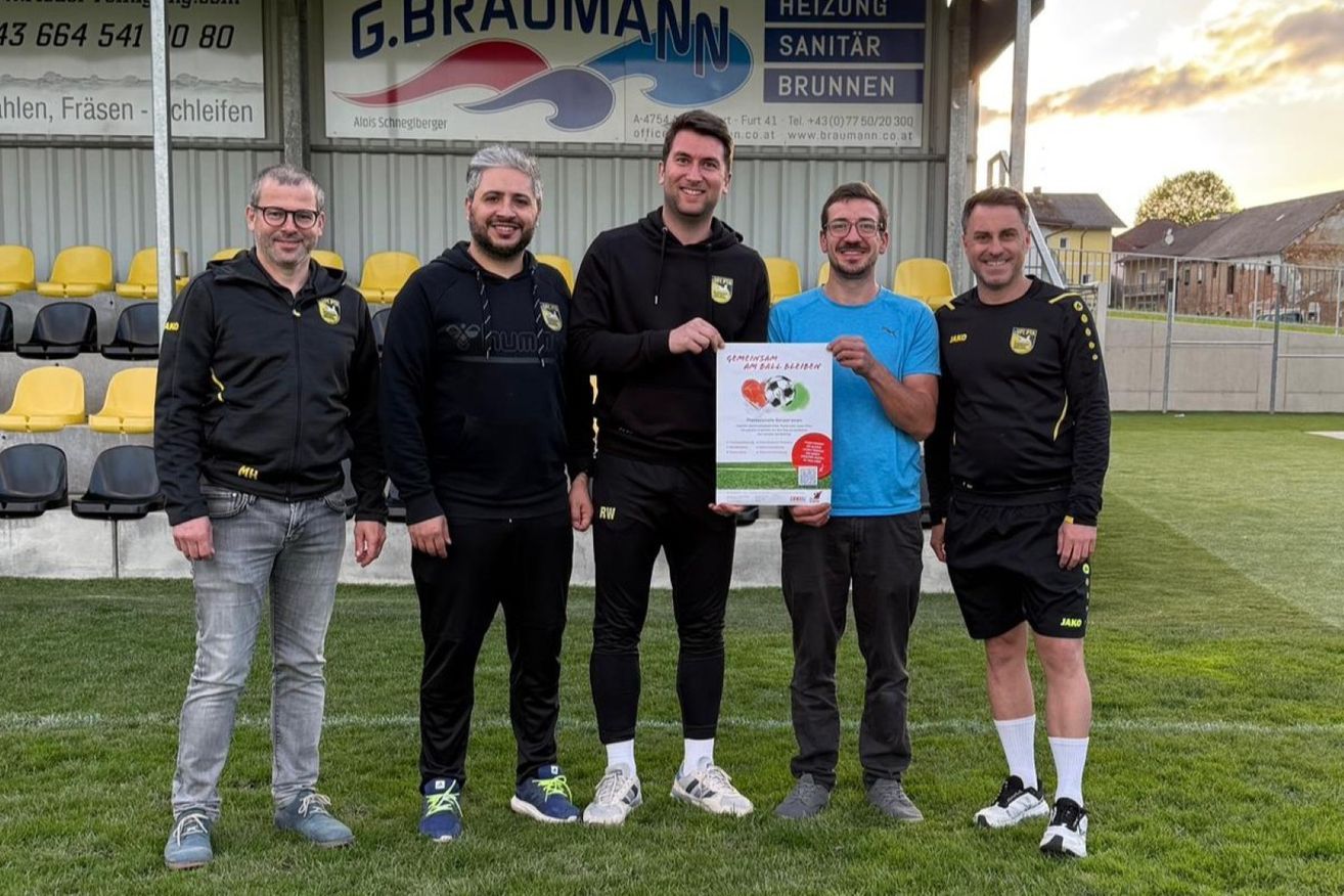 Fünf Männer stehen auf einem Rasenfeld. Einer hält ein Plakat. Sie tragen Sportbekleidung und Turnschuhe. Hinter ihnen zeigt ein Gebäude ein Schild mit dem Namen G.BRAUMANN und der Aufschrift Heizung Sanitär Brunnen.