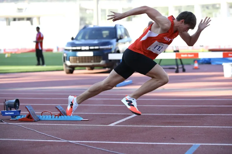 A runner with the number 112 on his shirt runs on a track with a blue car and a hurdle in the background.