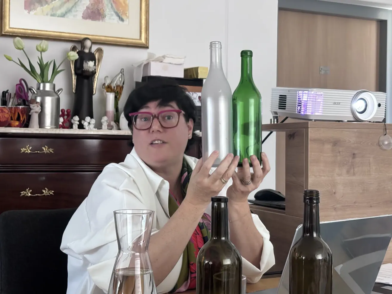 A woman in glasses holds two green bottles, one silver and one dark. She stands in a room with a shelf and a projector. A glass is filled with water nearby.