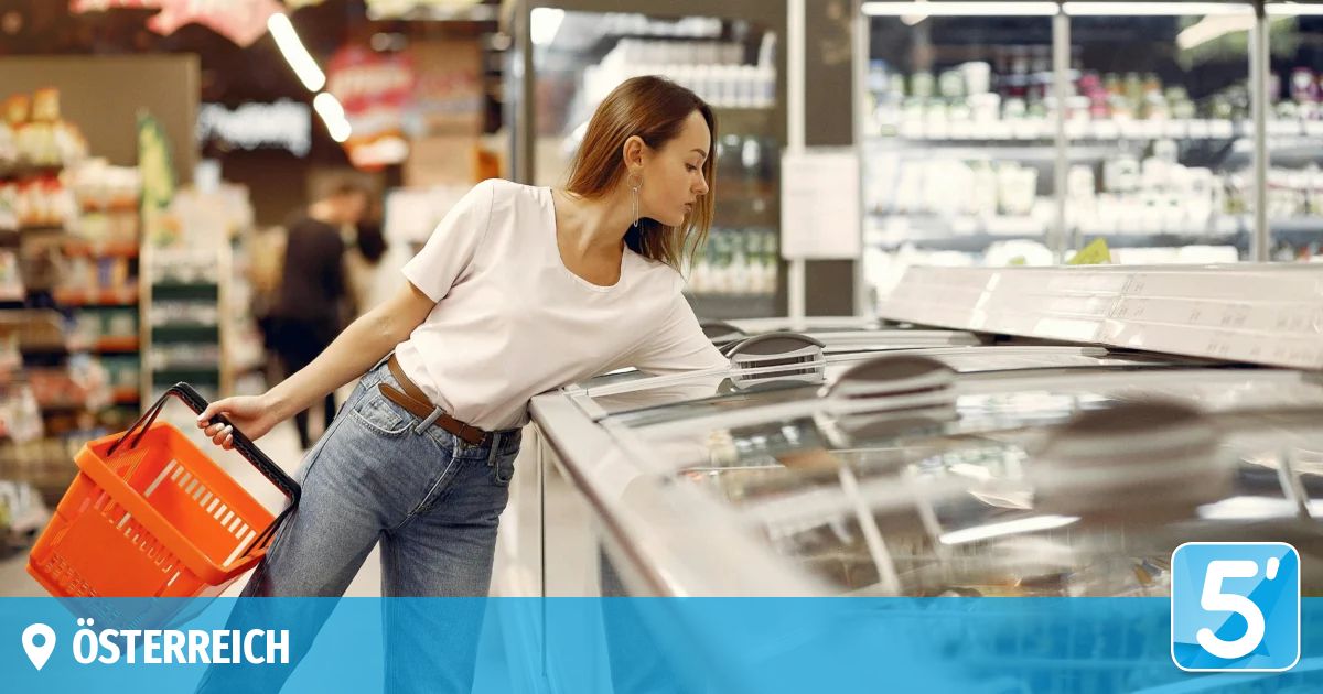 A woman in a white shirt and jeans is reaching into a freezer in a grocery store, holding a basket.