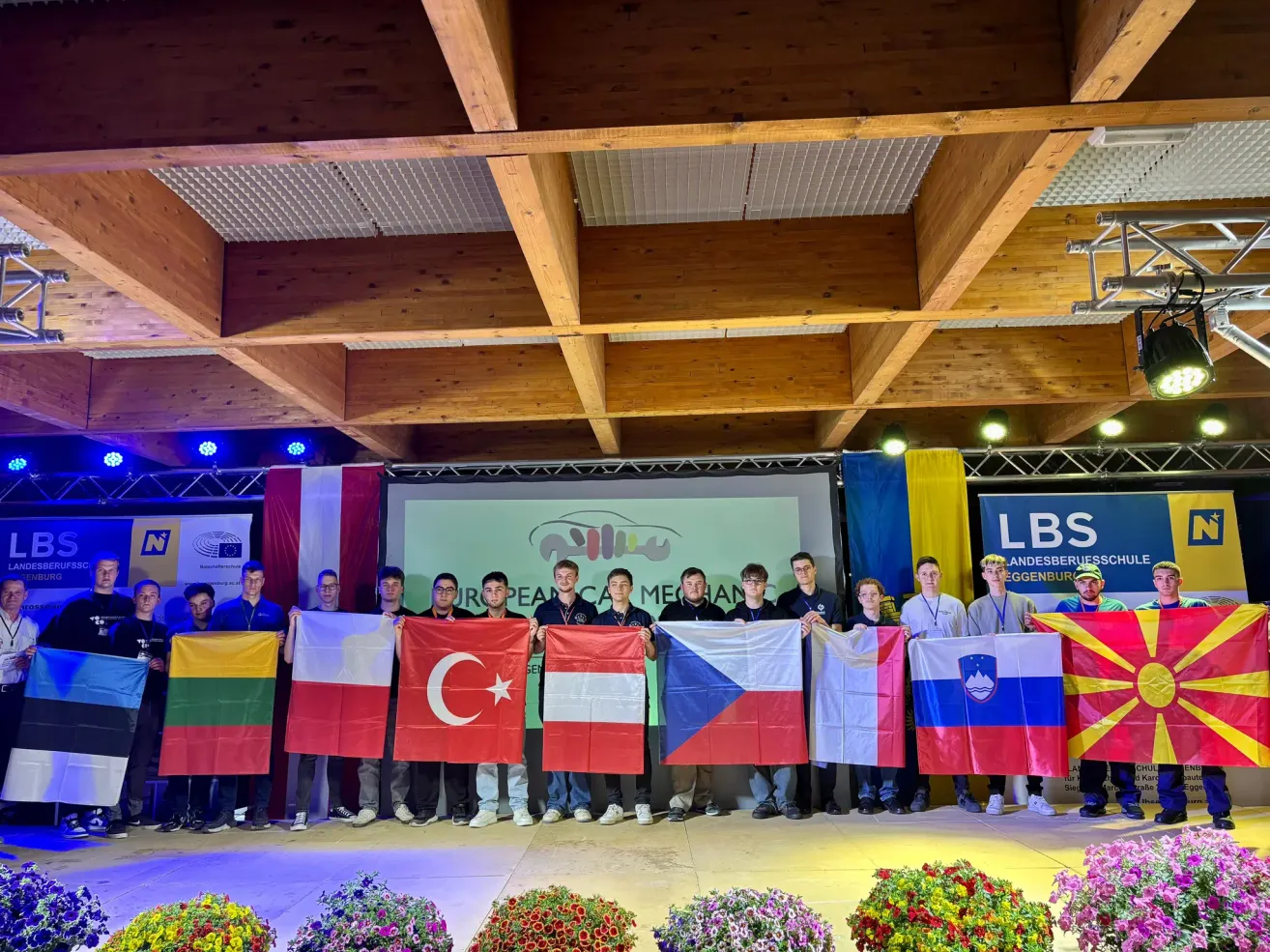 Group of individuals stand on stage holding flags from different countries. A projector screen is visible behind them.
