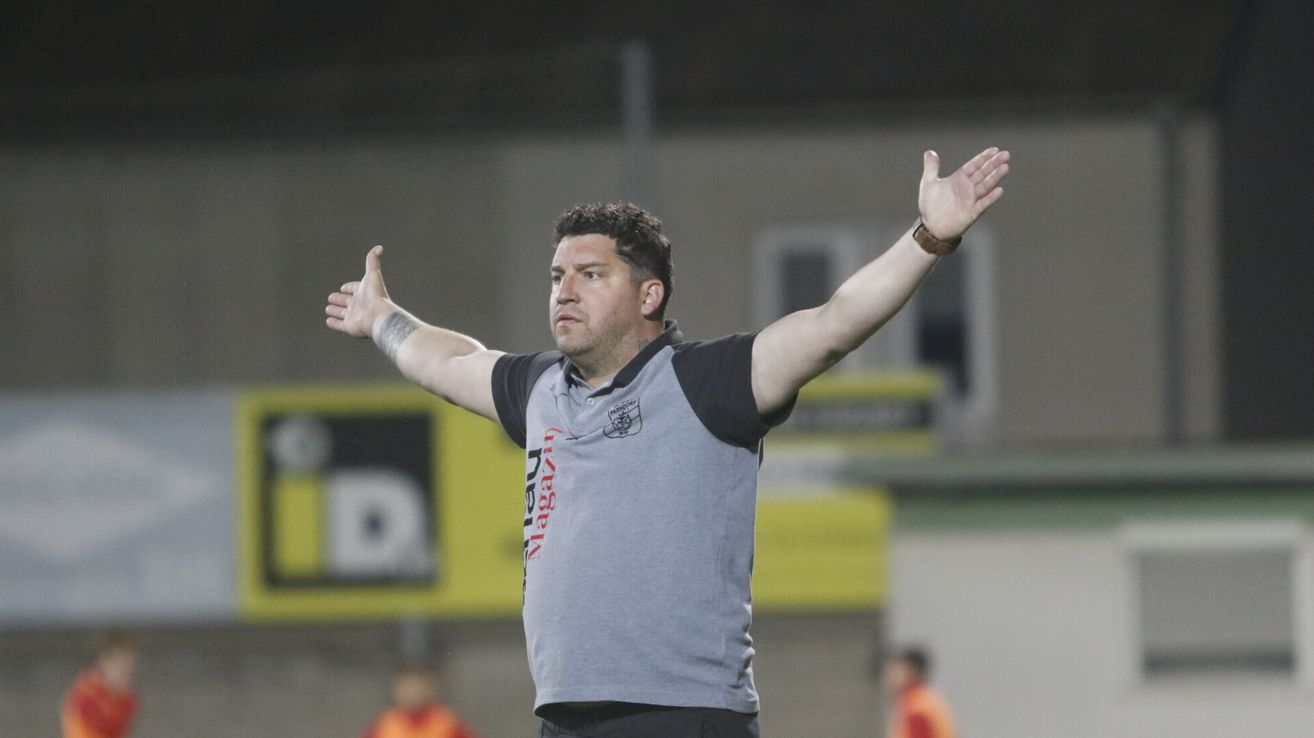 A man wearing a gray and black polo shirt stands on a field, gesturing with both arms raised. The word "Nizza" is visible on his shirt. Behind him, there is a blurred background of a yellow and white structure.