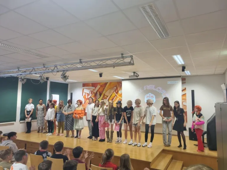 Eine Gruppe von Kindern in verschiedenen Kostümen steht auf einer Bühne vor einem Publikum, mit einer Projektionsleinwand im Hintergrund, die ein farbenfrohes Kunstwerk zeigt.