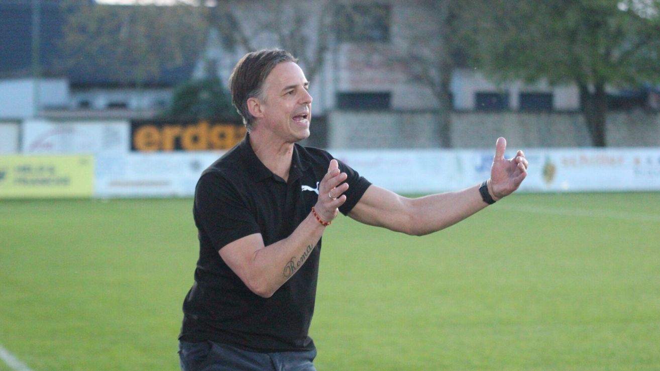 A man in a black polo shirt stands on a field, gesturing with his hands. He has a tattoo on his arm that reads 'Roma'. Behind him, a fence and buildings are visible.